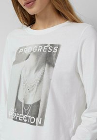 Biała bawełniana koszula z długim rękawem, ozdobiona graficznym nadrukiem naszyjników z napisem "PROGRES" oraz "nasza DOSKONAŁOŚĆ" w kolorze czarnym.