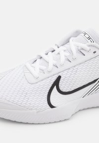Chaussure de sport blanche avec tige en mesh, arborant un logo swoosh noir et un design texturé. Comprend des lacets et une semelle en caoutchouc pour une meilleure adhérence.
