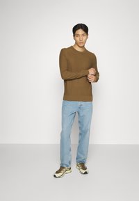 Brauner Strickpullover mit Rundhalsausschnitt, kombiniert mit hellblauen geraden Jeans und braunen sowie cremefarbenen Sneakers. Einfaches, sauberes Design.