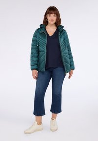 Žena stojící v zelenomodré prošívané bundě, námořnickém topu s výstřihem do V, zkrácených modrých džínách a bílých teniskách na pozadí jednoduchého bílého pozadí.