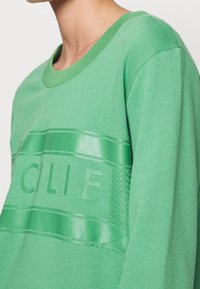 Il felpa verde presenta un design testurizzato con lettering in rilievo e accenti lisci. Collo a costine e texture in tessuto morbido.
