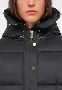 Zwarte pufferjas met een hoge kraag, gouden hardware accenten en een gewatteerd ontwerp. Heeft een ritssluiting aan de voorkant en drukknoopsluitingen.