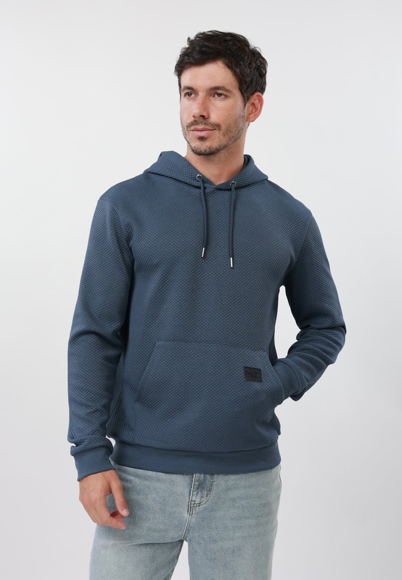 Homme aux cheveux courts et foncés portant un sweat à capuche bleu texturé et un jean bleu clair, debout avec une main dans la poche, regardant vers la droite.