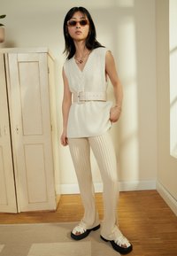 Top de malha creme sem mangas com decote em V, combinado com calças creme caneladas e sandálias chunky bicolores. Os acessórios incluem joias douradas e óculos de sol.