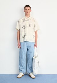 Korte mouwen, crèmekleurig shirt met zwarte abstracte prints, gecombineerd met lichtblauwe losse broek en witte tote bag, witte sneakers met beige accenten.