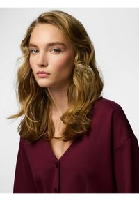 Bordo bluza s V-izrezom, velikim gumbima i širokim rukavima. Materijal izgleda glatko s mekanom teksturom.