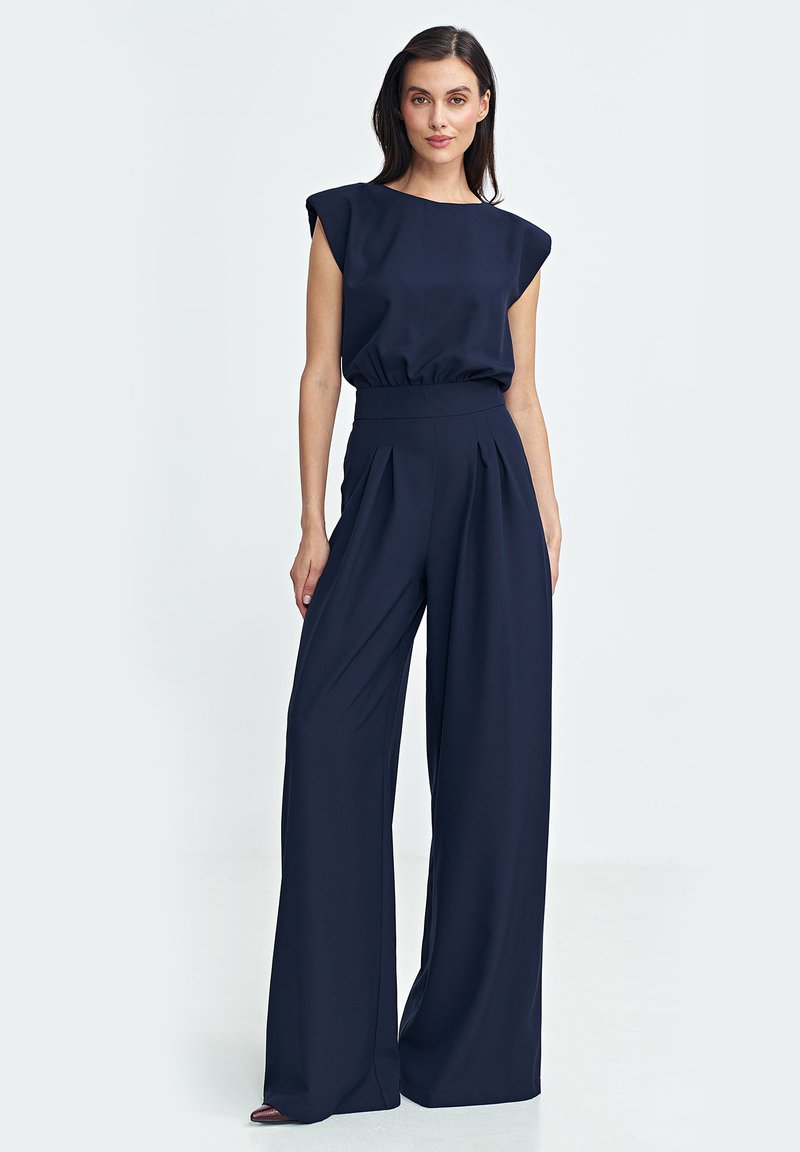 Granatowy kombinezon o szerokich nogawkach, z plisowanym przodem i krótkimi rękawami w stylu bufki. Wykonany z gładkiego, lejącego się materiału.