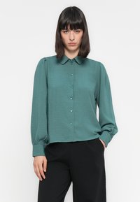 Τιρκουάζ πουκάμισο με γιακά, μακριά μανίκια και ελαφρώς φουσκωτές λεπτομέρειες, κατασκευασμένο από ελαφρύ, υφαντό ύφασμα. Συνδυασμένο με μαύρα παντελόνια.