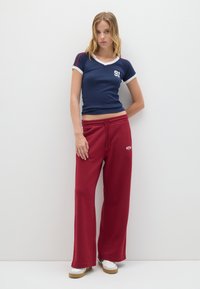 Maglietta blu navy con scollo a V e bordi bianchi, con il numero "91", abbinata a pantaloni tuta rossi larghi con vita elastica e logo. Scarpe bianche completano il look.