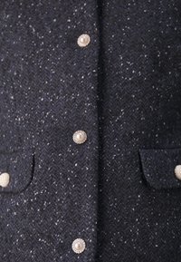 Tissu tweed bleu marine foncé avec des taches blanches et des boutons décoratifs en perle, avec de petites poches à rabat de chaque côté.