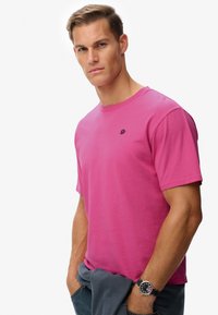 Camiseta de algodón de manga corta en color rosa, con un pequeño logo negro en el pecho. El modelo lleva una chaqueta gris colgada del brazo.