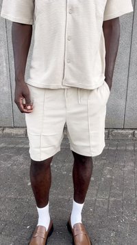 Beige short-sleeve, textured shirt and matching beige shorts with drawstring. Brown loafers worn with white socks, standing on a paved surface.