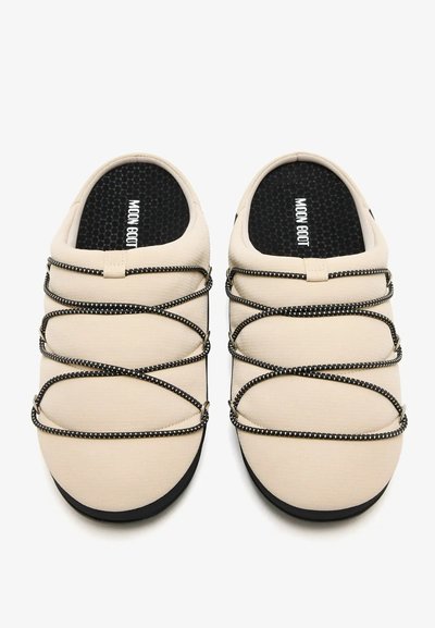 Chaussures slip-on beiges avec semelles intérieures rembourrées noires marquées "MOON BOOT", avec des lacets élastiques noirs croisés sur le dessus.
