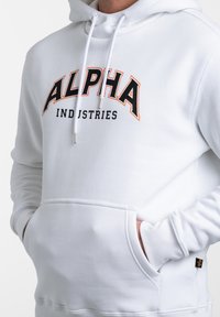 Sudadera blanca con un bolsillo tipo canguro, cordones y un gran logo de "ALPHA INDUSTRIES" en negro y naranja en el pecho.