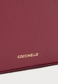 Borsa in pelle strutturata color bordeaux con logo dorato "Coccinelle". Caratterizzata da una finitura liscia, forma rettangolare compatta e cuciture robuste.