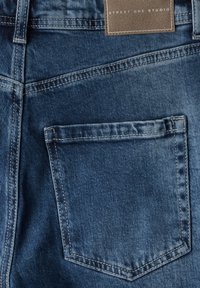 Jeans de denim azul oscuro con un bolsillo trasero visible y detalles de costura. Una etiqueta de tela dice "STREET ONE STUDIO". Textura suave.