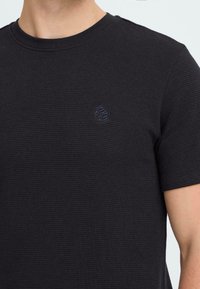 Fekete, textúrájú, rövid ujjú póló, kerek nyakkal és kis logó részlettel a mellkason. Az anyag rácsos mintázatú.