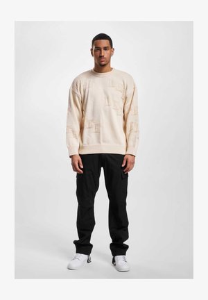 Pull en tricot beige avec des motifs en relief, col rond et épaules tombantes, associé à un pantalon cargo noir et des baskets blanches.