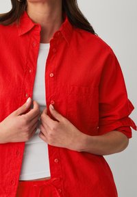 Czerwona koszula button-up z lekkiego materiału, z kieszonką, podwiniętymi rękawami i kołnierzykiem, noszona na białej dopasowanej bluzce.