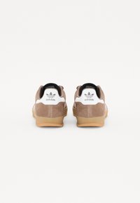 Hnědé semišové tenisky s bílými akcenty, gumovou podrážkou a černým lemováním. Na patě a po stranách jsou logo Adidas a pruhy.