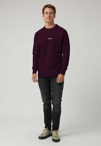 Hombre vistiendo una sudadera morada oscura con texto blanco, jeans negros y zapatillas beige, de pie contra un fondo claro y liso.