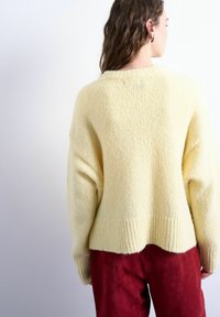 Femme aux longs cheveux bruns portant un pull en maille jaune pâle surdimensionné et un pantalon rouge taille haute, vue de dos sur un fond clair uni.