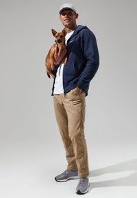 Navy zip-up hoodie, white t-shirt, tan pants, and gray sneakers. Holding a small brown dog. Simple, casual design with neutral tones.