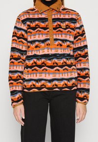 Bluza polarowa z pomarańczowymi, różowymi i czarnymi wzorami plemiennymi; posiada brązowy kołnierz na zatrzaski i detal z kieszenią z przodu.