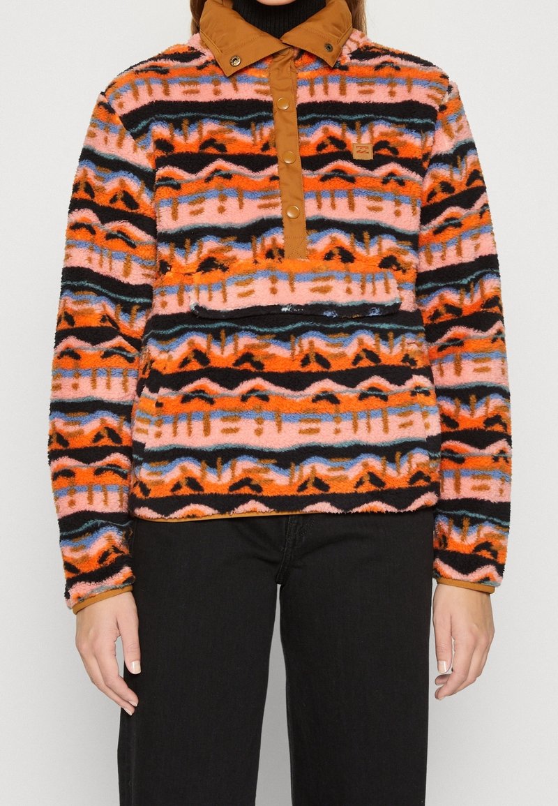 Bluza polarowa z pomarańczowymi, różowymi i czarnymi wzorami plemiennymi; posiada brązowy kołnierz na zatrzaski i detal z kieszenią z przodu.