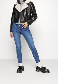 Chaqueta corta de cuero negro con cuello de borrego, combinada con jeans ajustados azules y zapatos negros con detalle de borlas.