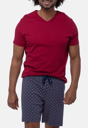 Mand iført en rød T-shirt med korte ærmer og V-hals samt marineblå mønstrede shorts med snøre i taljen, smilende.