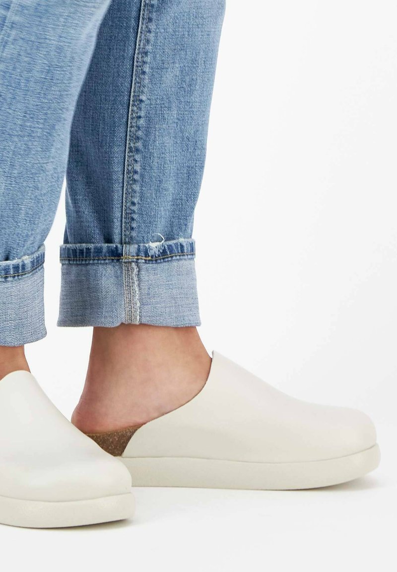 Chaussures blanches à enfiler avec une tige lisse, accent en liège au talon, associées à un jean bleu clair retroussé.