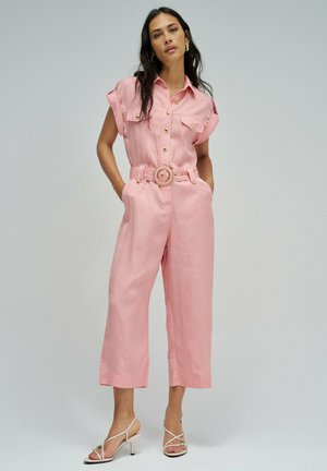 Mujer de cabello largo y oscuro, vestida con un mono rosa con cinturón, mangas remangadas y sandalias beige de tacón alto con tiras, de pie con las manos en los bolsillos.