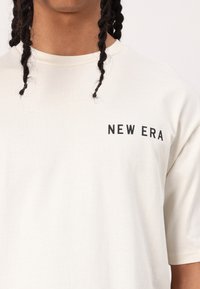 Cremefarvet bomulds T-shirt med rund hals og korte ærmer, prydet med et sort "NEW ERA" logo på venstre brystområde.