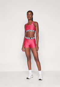 Roter sportlicher Sport-BH und Shorts mit schwarzem "UNDER ARMOUR"-Bund. Glattes, dehnbares Material. Das Model steht vor einem neutralen Hintergrund.