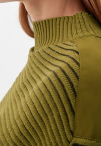 Pull-over vert olive avec une texture côtelée, présentant des rayures verticales et un col haut. Le tissu lisse sur les épaules ajoute un contraste.