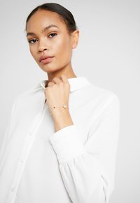 Camisa blanca de botones con puños enrollados, combinada con una delicada pulsera de oro que presenta un pendant circular y un pequeño acento de cuentas.