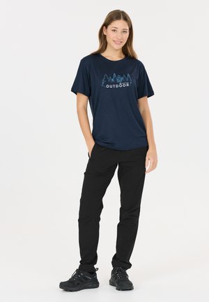 Frau steht, trägt ein marineblaues T-Shirt mit Baumgrafik und dem Schriftzug „OUTDOOR“, schwarze Hose und schwarze Wanderschuhe vor weißem Hintergrund.