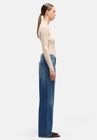 Jasnobeżowa bluzka z długim rękawem w połączeniu z niebieskimi dżinsami o wysokim stanie i luźnym krojem. Dżinsy mają tylne kieszenie i prostą nogawkę.