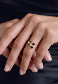 Gold ring featuring black and clear square accents, with a geometric design. Smooth texture, set against a hand with natural nails.