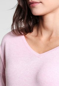 Vaaleanpunainen v-aukkosweater, pehmeä pinta, ribattu kaulus ja rento malli. Lähikuva, joka tuo esille kankaan yksityiskohdat ja värin.