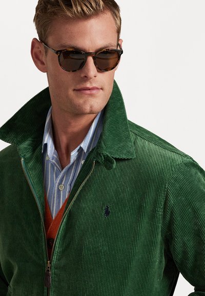 Giacca in corduroy verde con chiusura a zip, colletto alto e logo blu del polo. Abbinata a una camicia a righe blu e un cardigan arancione. Occhiali da sole marroni.