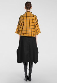Musztardowożółty dzianinowy top w czarną kratę, z szerokimi rękawami i wysokim kołnierzem, zestawiony z długą czarną spódnicą i błyszczącymi czarnymi botkami.