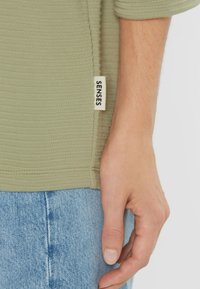 Olive green textured top with ribbed fabric, side seam label reading "SENSES," paired with light blue denim. Close-up on hand resting on fabric.