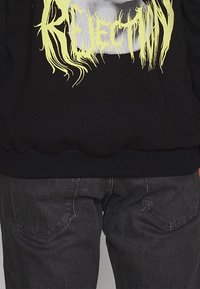 Felpa nera con la scritta "REJECTION" in lettere gialle e seghettate, abbinata a jeans in denim grigio scuro con tasche posteriori e una texture leggermente consumata.