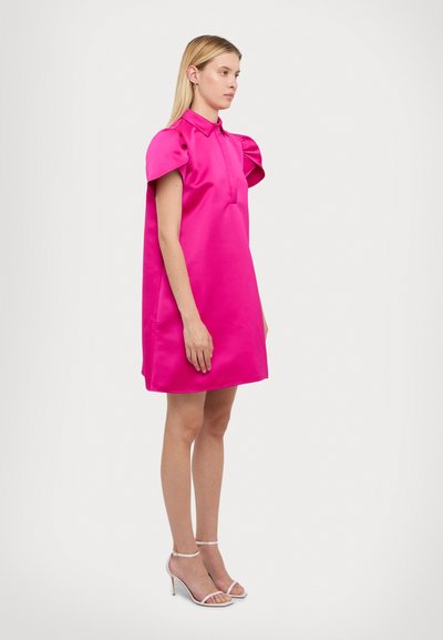 Robe en satin fuchsia avec un col à revers et des manches bouffantes, présentant une silhouette droite et un ourlet au genou. Associée à des talons blancs à lanières.