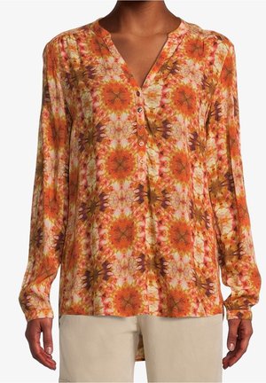 Blouse à manches longues avec motif floral circulaire orange, jaune et marron et patte de boutonnage partielle portée avec un pantalon beige clair.