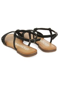 Sandalias trenzadas negras con suela plana y plantilla marrón. Correa ajustable en el tobillo con hebilla de metal, diseño minimalista para un uso fácil.
