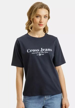 Kobieta o falowanych blond włosach, ubrana w granatowy t-shirt "Cross Jeans Sound of the Sea" oraz jasnoniebieskie dżinsy, stojąca na białym tle.