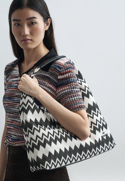 Young woman with long dark hair wearing a multicolored striped top, holding a large black-and-white zigzag patterned shoulder bag.